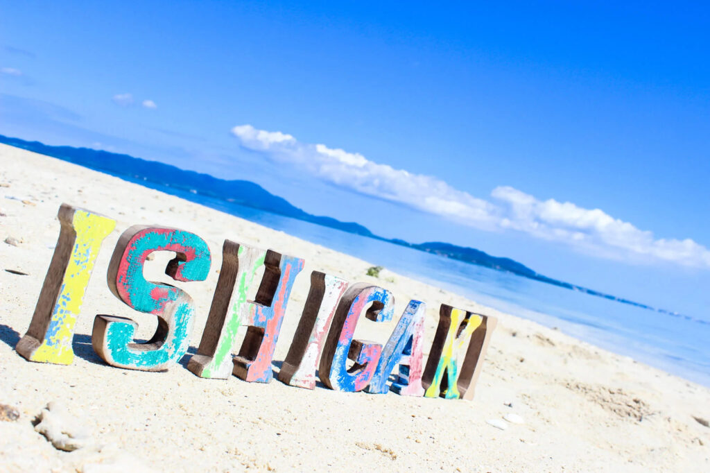 砂浜に「ISHIGAKI」と書かれた文字ブロックを置いています。青い空にカラフルな文字が映えています